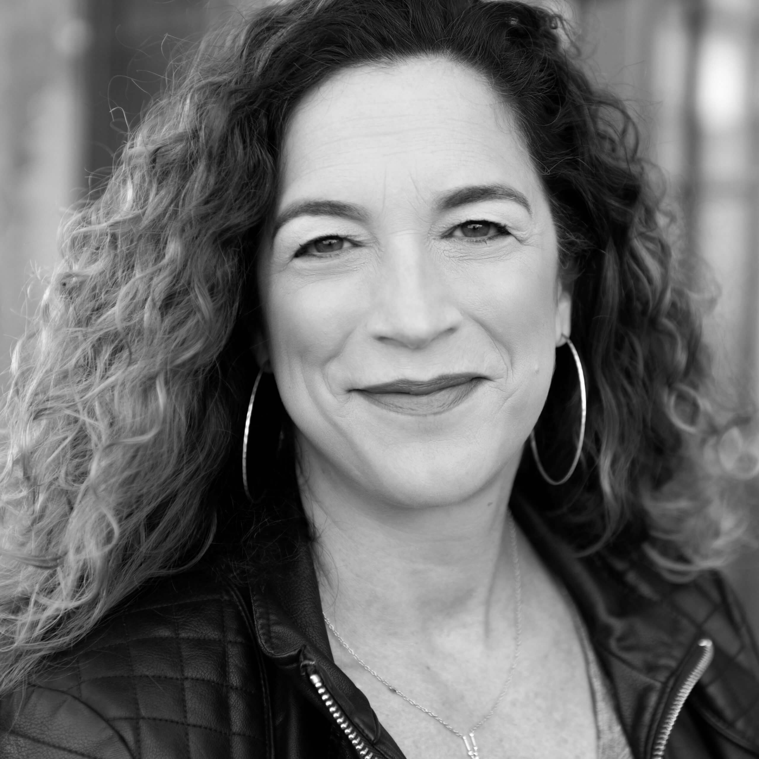 Kristi Jacobson slightly smiling and looking directly at the camera. She has medium curly hair and wears a jacket, thin hoop earrings, and a necklace. Portrait in black and white.