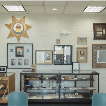 Still from Sanctuary. A wall is full of framed pictures, metals, badges and other items honoring a Sherrif's office. Below the hanging items are two display cases full of other items such as helmets, guns, etc.