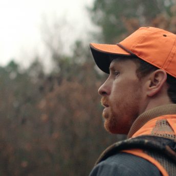 Still from One Shot One Kill. A man standing outside looks off camera, he is wearing an orange vest and hat.