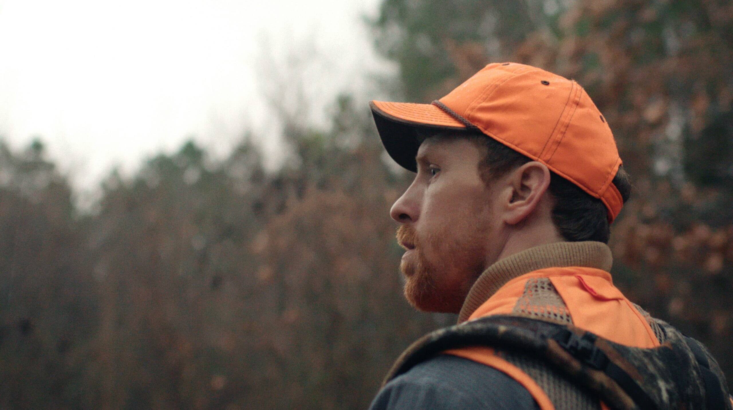 Still from One Shot One Kill. A man standing outside looks off camera, he is wearing an orange vest and hat.