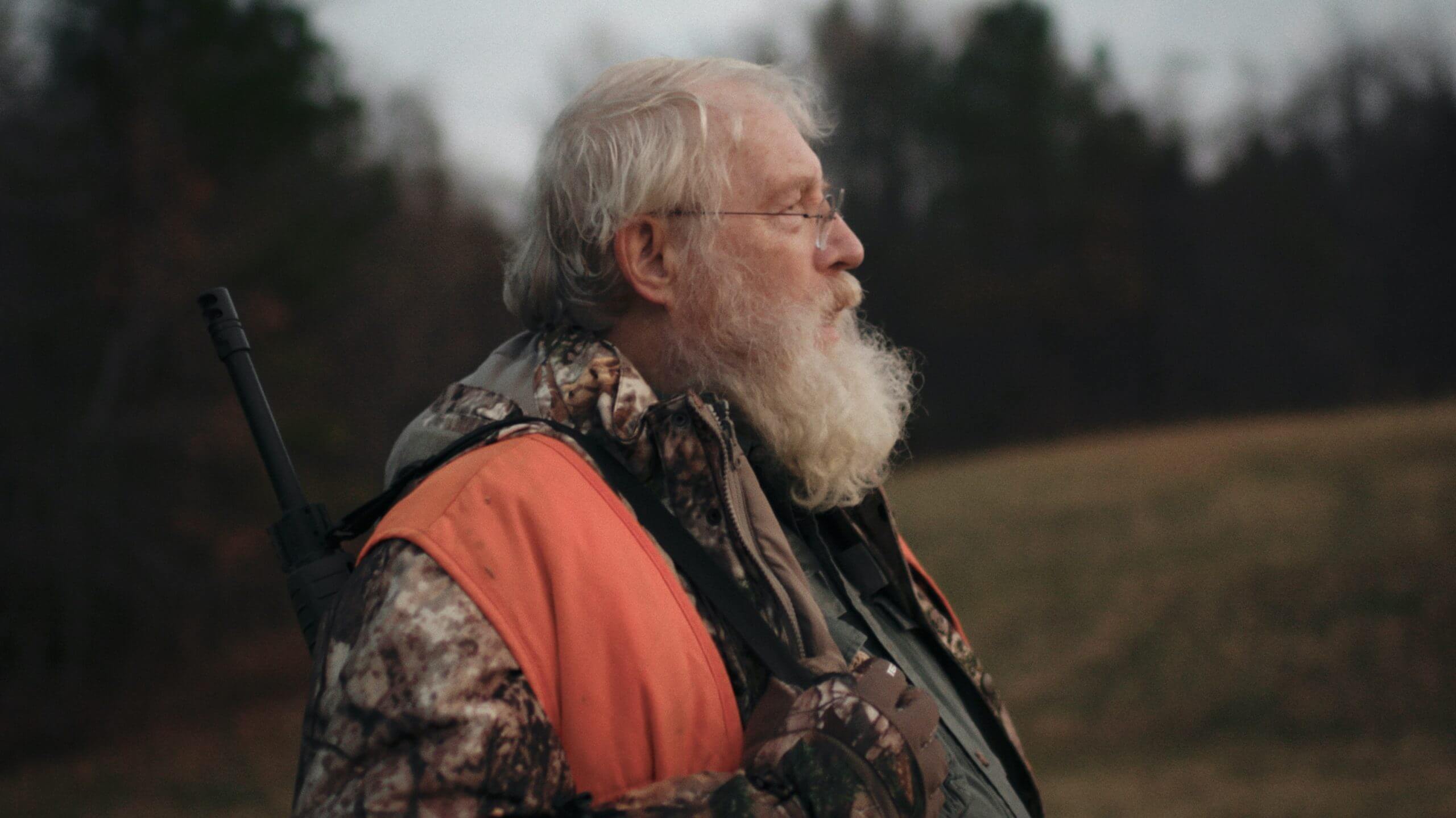 Still from One Shot One Kill. A man is standing outside, wearing a camoflauge jacket and orange vest with a gun strapped over his right shoulder.