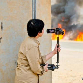A woman is filming a fire from behind the corner of a building. There is a copyright symbol with the text "2021 Guillame Briquet" on the bottom right corner.