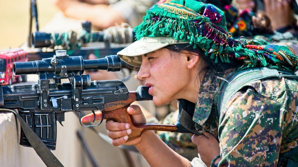 A woman in a military uniform and colorful headdress gazes into the scope of a gun leaning on a cement barrier. There are other people behind her doing the same thing.