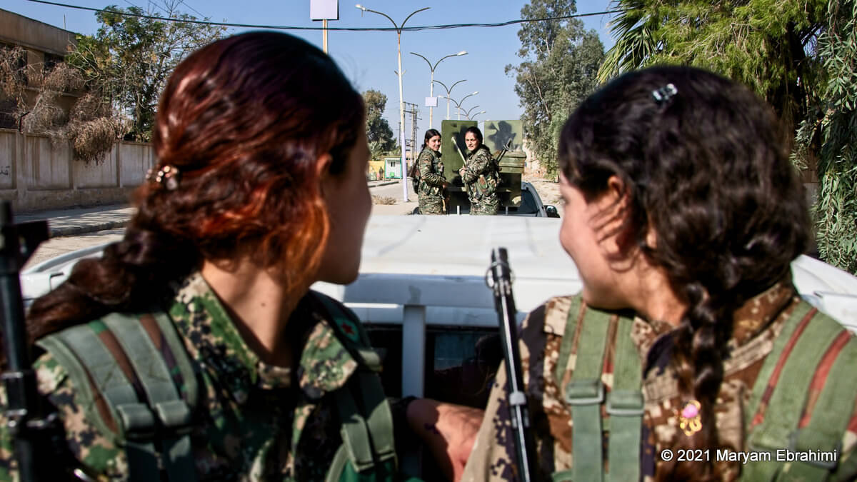 Two women in military uniforms look behind them towards two more women in uniform standing in the back of a truck.