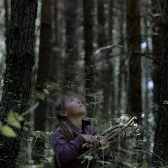 Still from El Eco. A young girl stands in the center of a forest. She is facing to the side and is looking up. She holds a bundle of small branches in her arms.