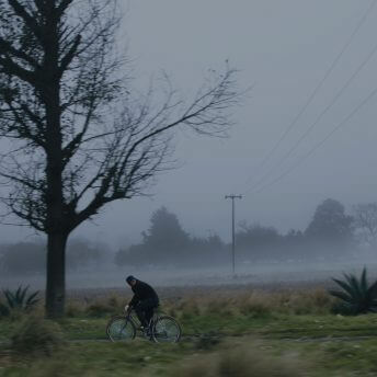 Still from Tempestad. A man rides a bicycle down a rural street under a cloudy sky.