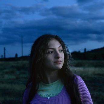 Still from Tempestad. A girl stands in front of the camera and looks away to the left. She is in the middle of a grassy plain with dark clouds in the sky.