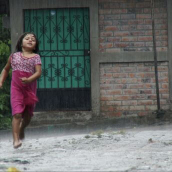 Still from The Tiniest Place. A little girl running down the street in the rain. She is barefoot and wearing a pink dress.