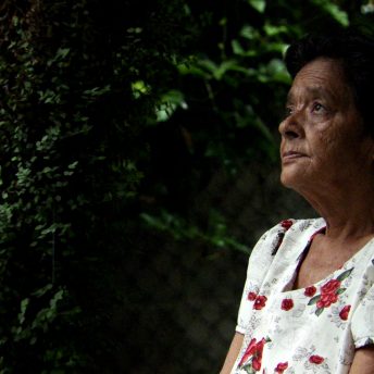Still from the Tiniest place. A side shot of an old woman looking to something offscreen. She wears a white t-shirt with red flowers patterning it.