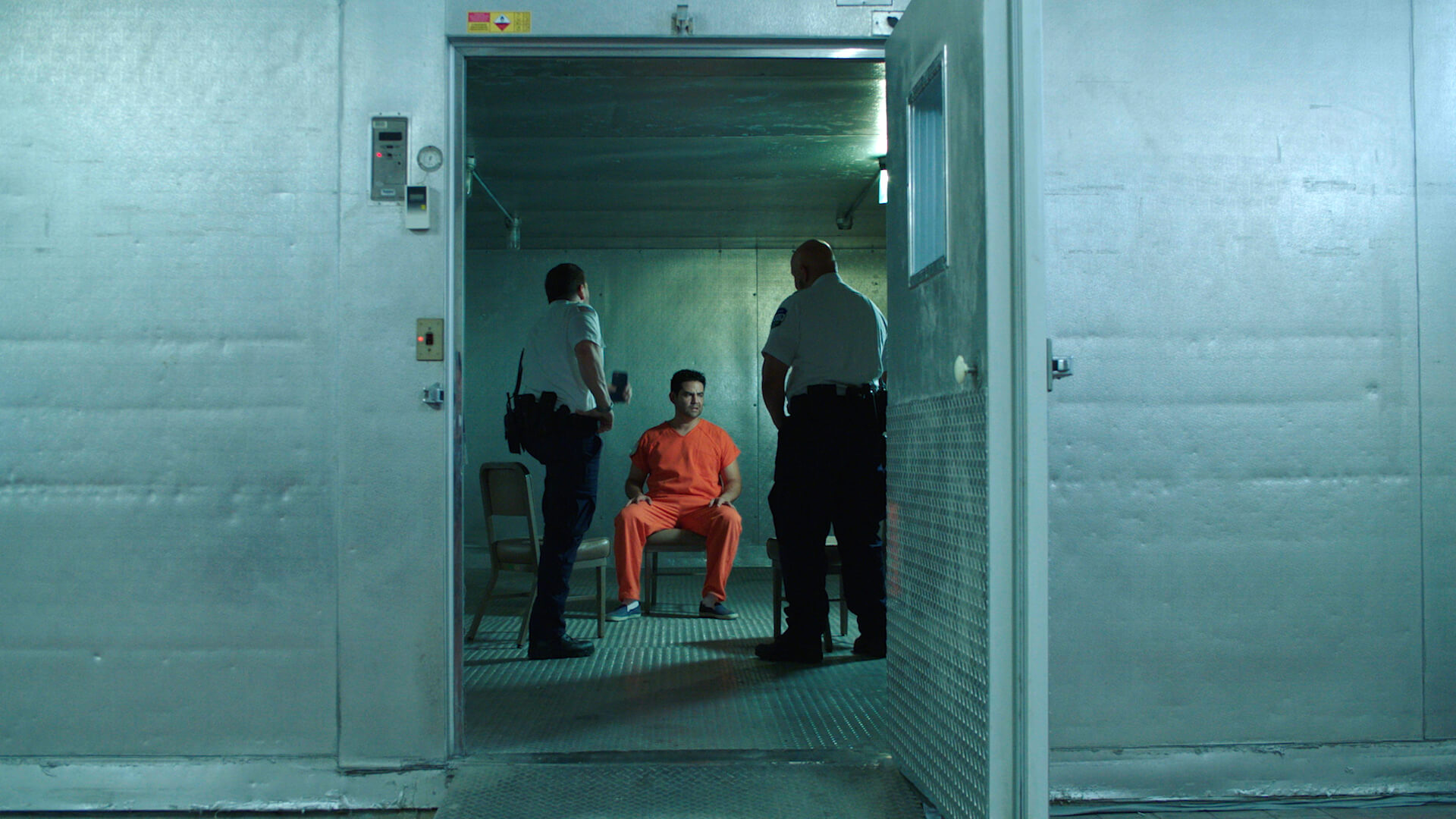 Two prison guards talk to a man in a prison uniform. They are standing in front of him, while he is sitting on a chair.