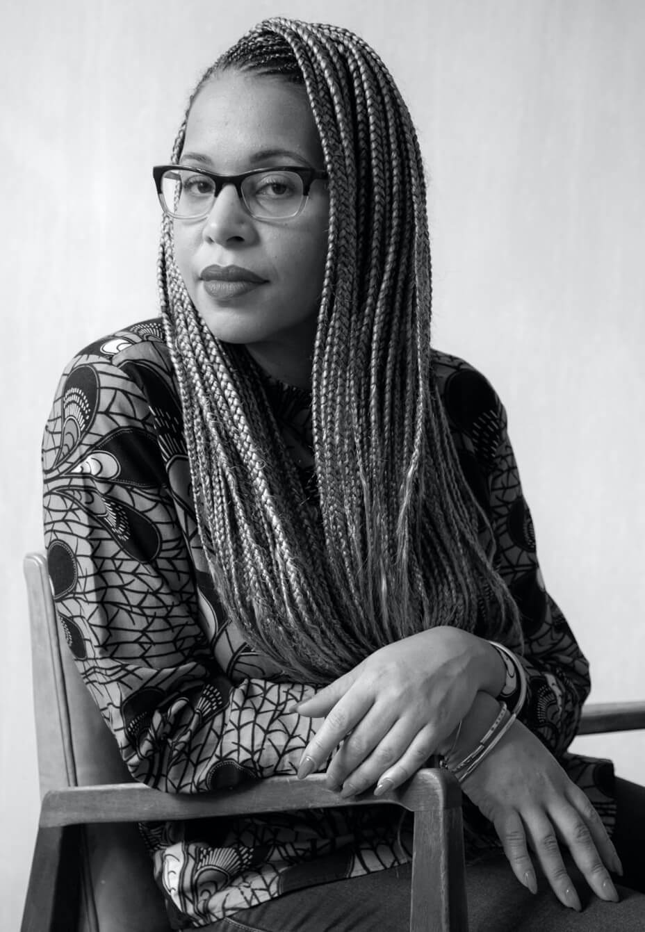 Loira Limbal is sitting in a chair, she looks at the camera with her head slightly turned. Loira wears lipstick and her hair is braided. Portrait in black and white.