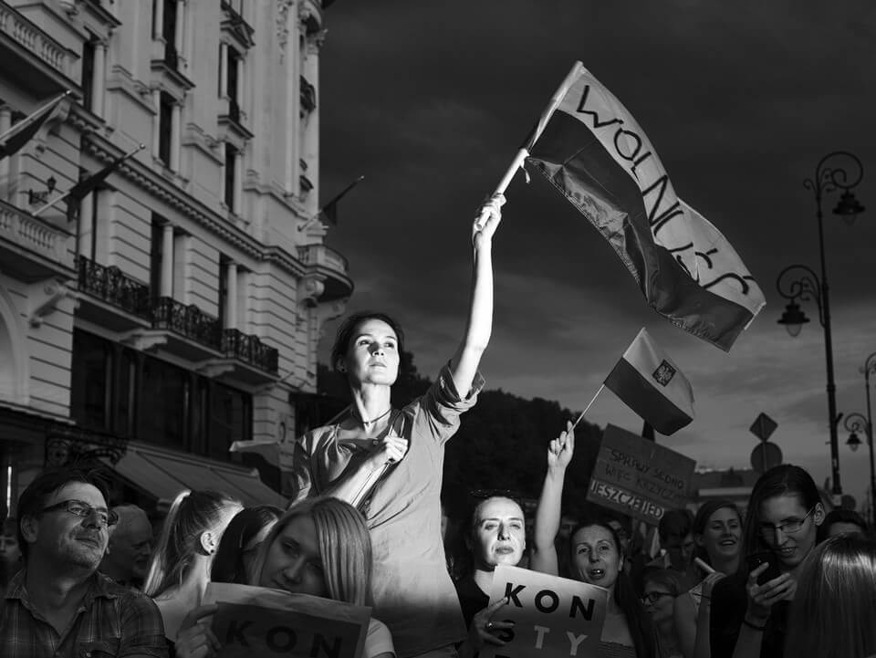 Black and white still from Freedom Highway. A woman is holding up a flag above her head. She is illuminated and the focus of the photo, among a crowd of people who are also holding signs and flags.