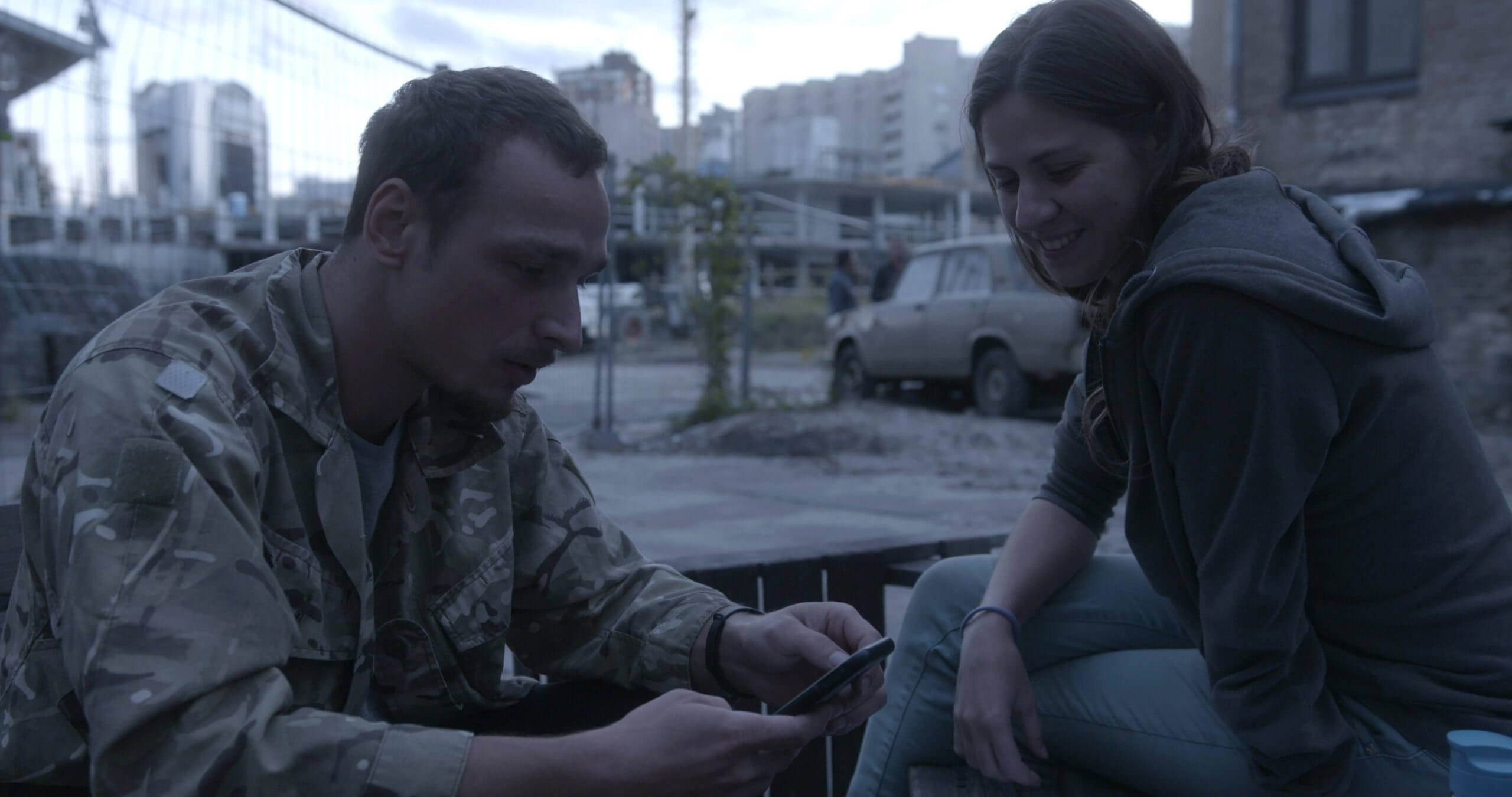 Still from The Hamlet Syndrome. A man and a woman look at a cellphone. Both are sitting. The man wears a camo jacket, the woman is dressed in jeans and a hoodie. The background is a cityscape.