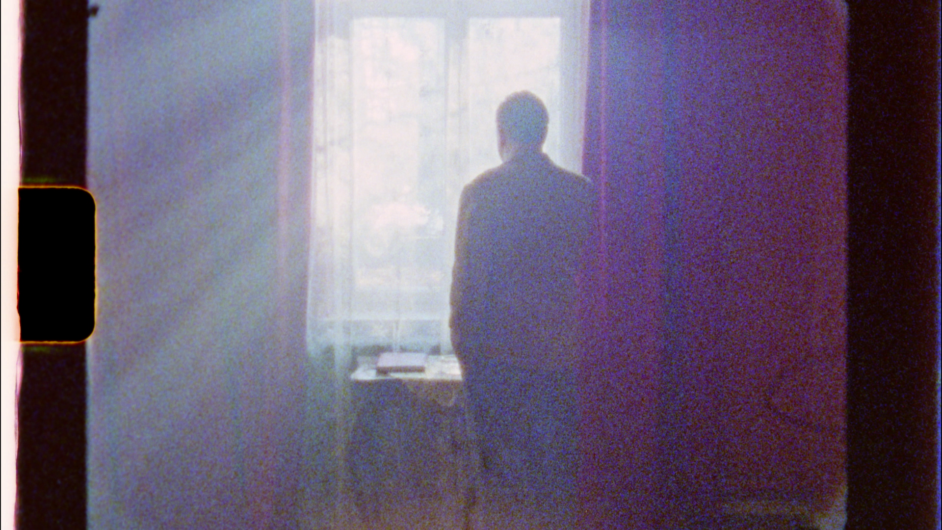 Still from The Prince and the Dybbuk. A man is standing in front of a brightly-lit window. The photo appears to be a copy of an analog film still. The composition is grainy and overexposed.