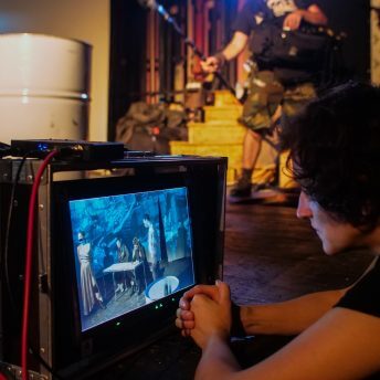 Production still. Elwira Niewiera watches a monitor of a scene that she is shooting. A man holding a boom mic is seen in the background.