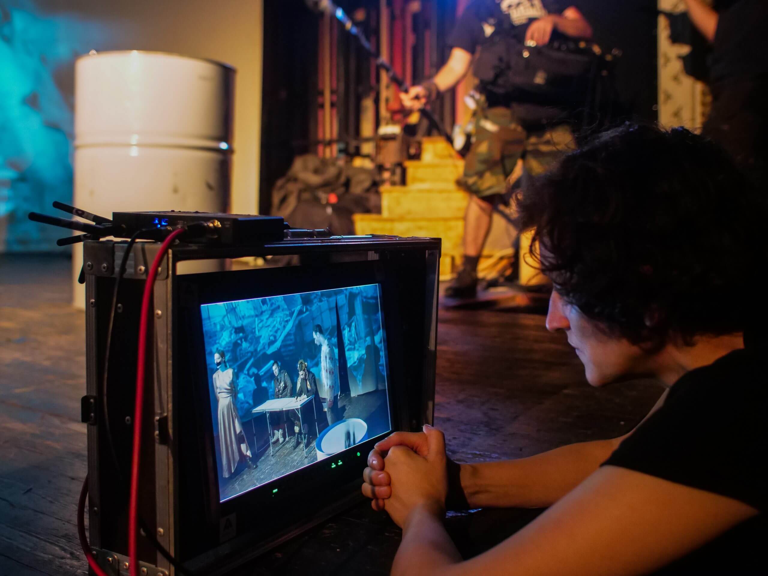 Production still. Elwira Niewiera watches a monitor of a scene that she is shooting. A man holding a boom mic is seen in the background.