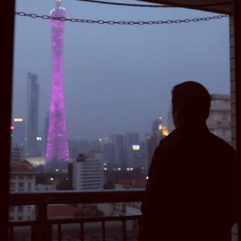 Still from Complicit, a film directed by Jialing Zhang. A man is pictured from behind as a dark silhouette against a twilight sky and cityscape dominated by a tall tower of pink lights.