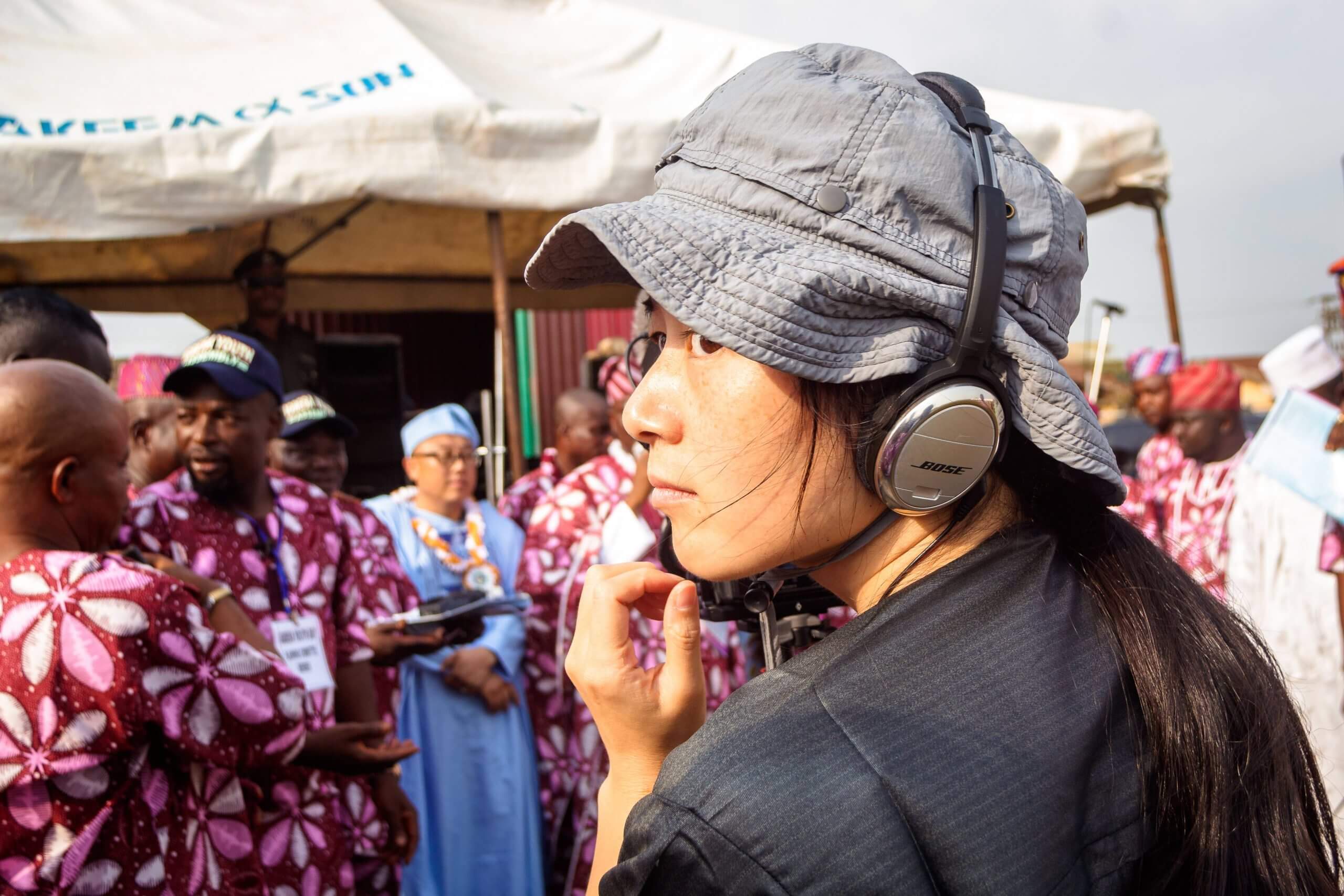 Production still of Jialing Zhang. She is wearing a wide-brimmed hat and headphones. She is pictured from the back, looking to the side and out of the frame. Behind her, slightly focused, there is a group of men in brightly-colored and patterned clothes.