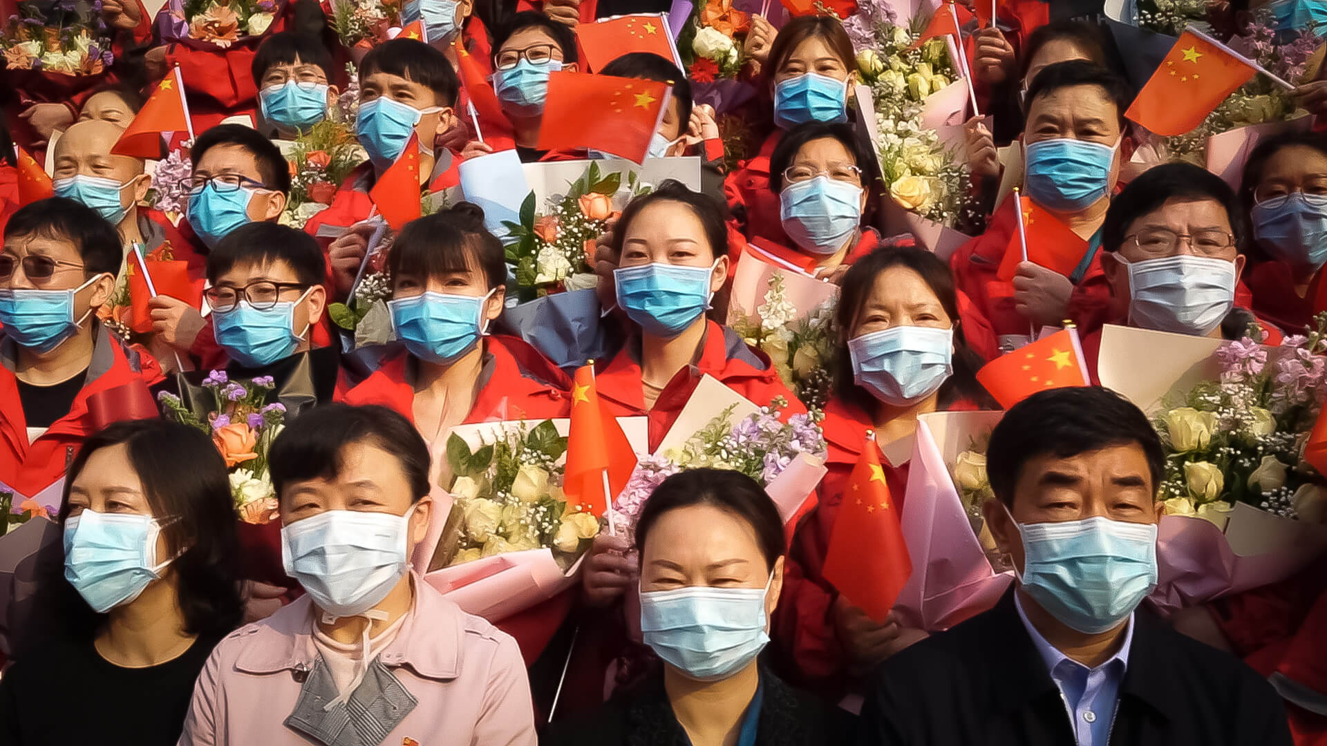 Still from In the Same Breath, a film produced by Jialing Zhang. Group portrait of Chinese men and women who are standing shoulder to shoulder in stadium-style rows. They are holding Chinese flags and some women are holding bouquets of flowers. They are all wearing blue surgical masks.