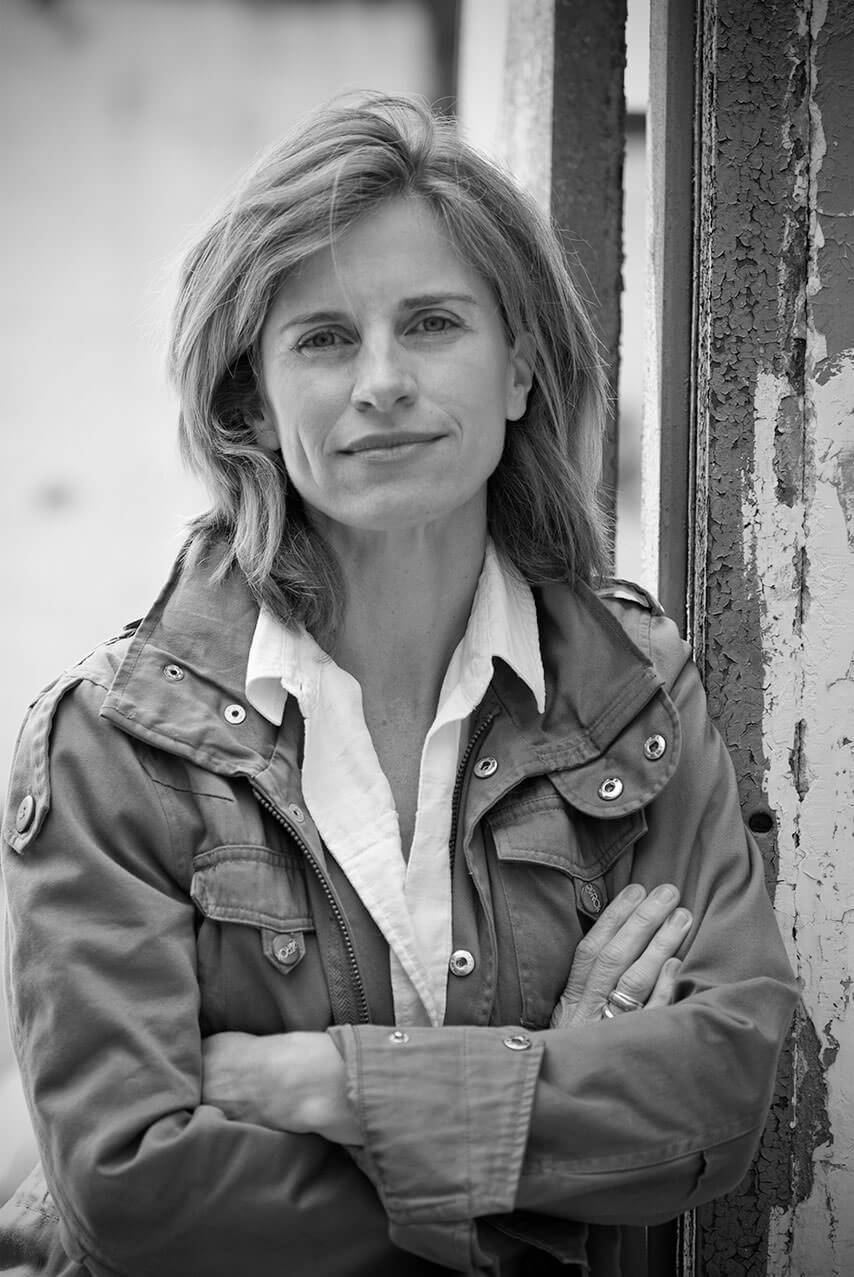 Black and white headshot of Holly Morris from the waist up with relaxed crossed arms and closed mouth, wearing a cargo jacket