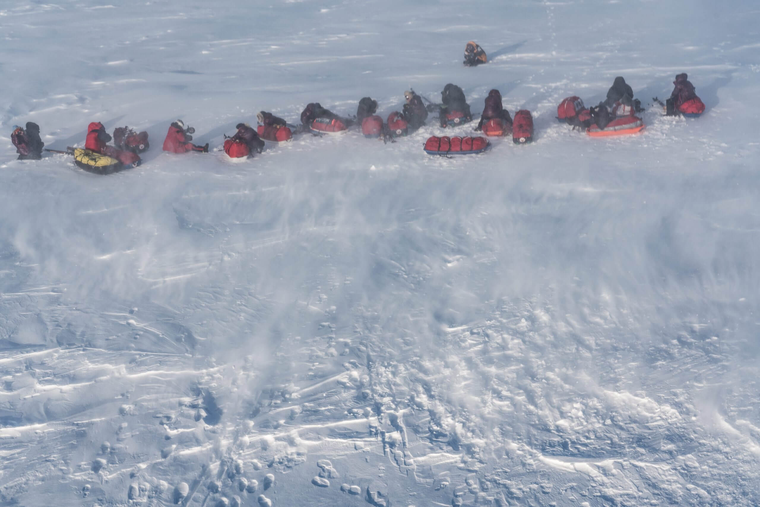 Bird's eye view of approximately 20 people in winter gear sitting and laying in a horizontal line across the snow.