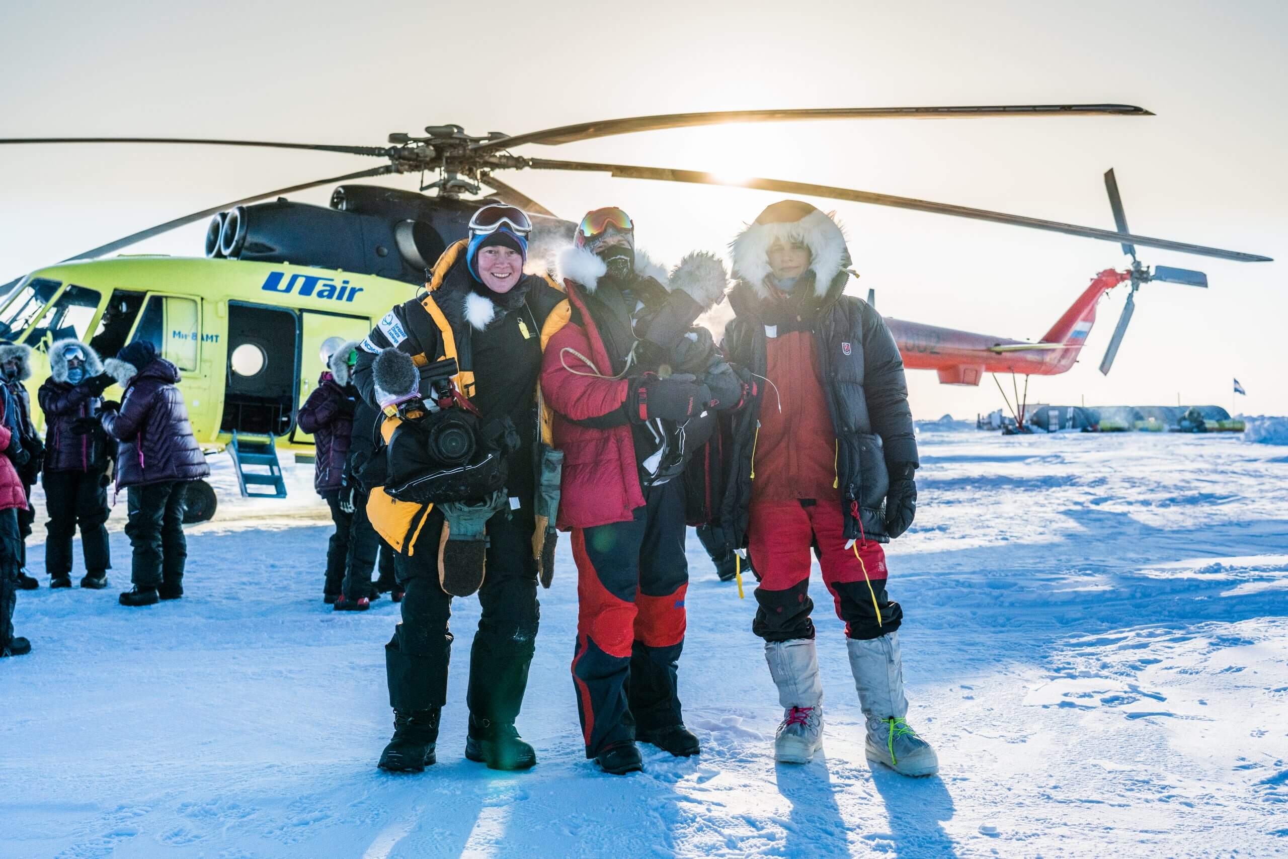 Three women in winter gear in the snow in front of a grounded helicopter, facing forward with the sun shining behind them.