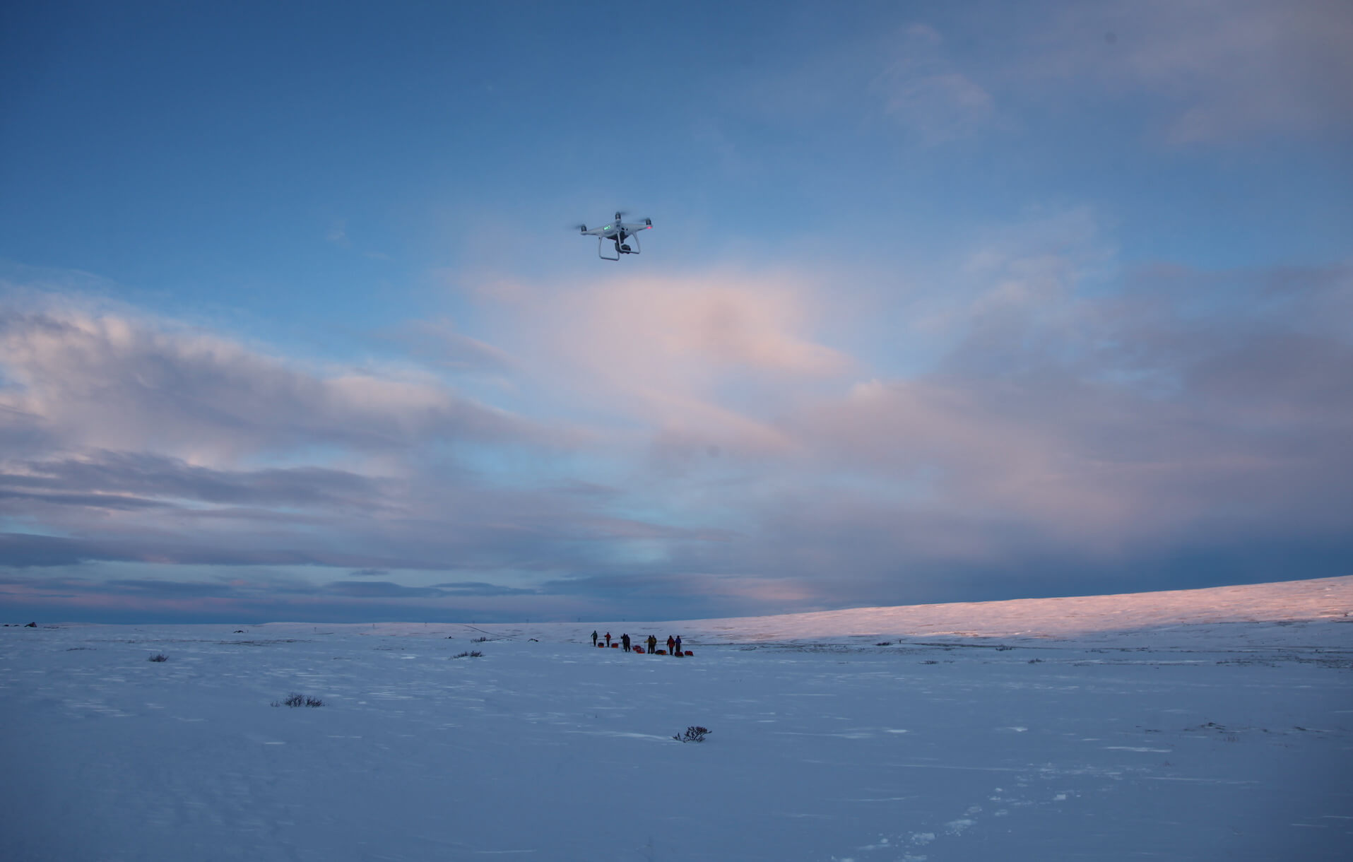 Snowy flat landscape with a drone flying in the peach and blue sky with some clouds and a group of people in the distance.