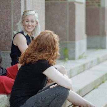 Three women sitting on the steps of a building are talking with each other. One of them, with red hair, gives her back to the camera to face one of the women.