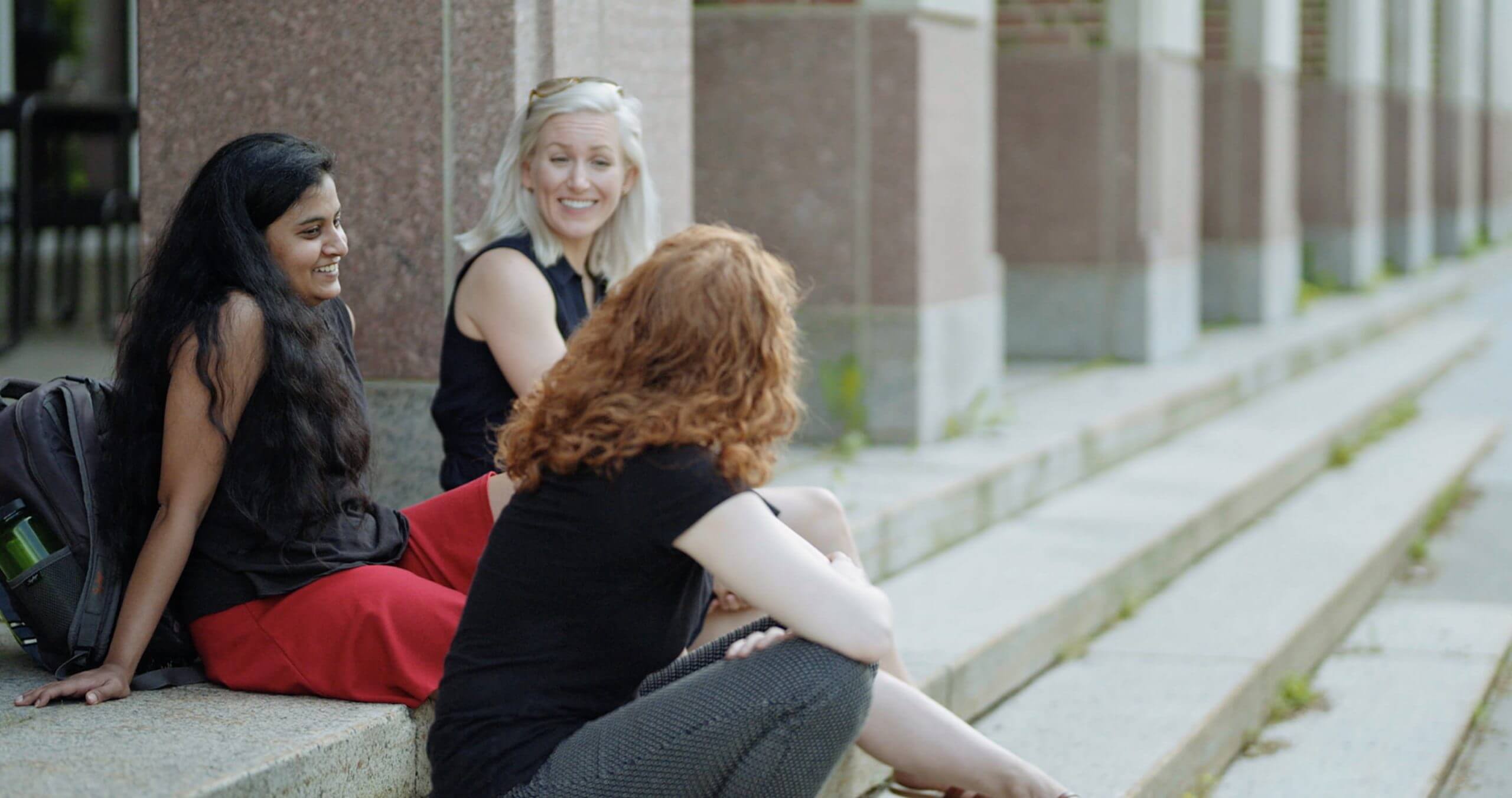 Three women sitting on the steps of a building are talking with each other. One of them, with red hair, gives her back to the camera to face one of the women.