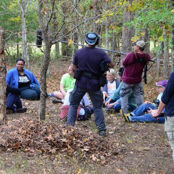 Production shot of Acts of Reparation. The crew is filming a group of people sitting on the ground in the woods.