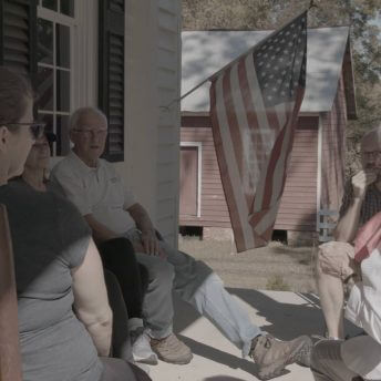Still from Acts of Reparation. Shot of a group of white people sat in chairs in the porch of a house with a US flag in the back. It is sunny.