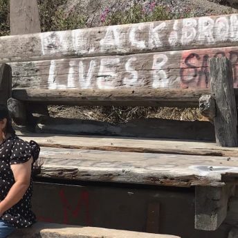 Still from Acts of Reparation. Medium shot of a woman wearing a polka dot shirt, sitting in front of a woody bench that has written: "Black & brown lives r important". She looks away from the camera.
