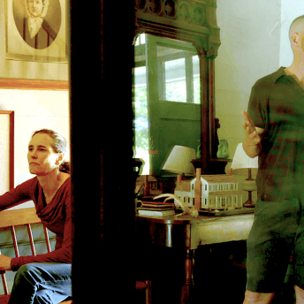 Still from Acts of Reparation. Shot of a woman sat in a chair and the reflection of Macky Alston standing while talking and gesticulating with his arms, they are both inside of a house.