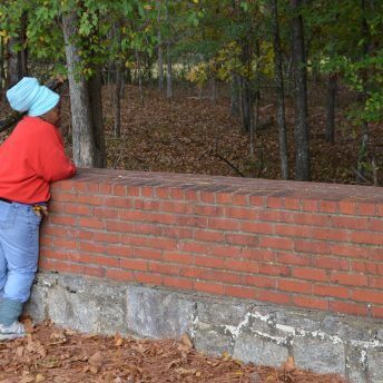 Still from Acts of Reparation. Full shot of a woman standing while leaning on a short wall in the forest. She wears a red hoodie and light blue jeans.
