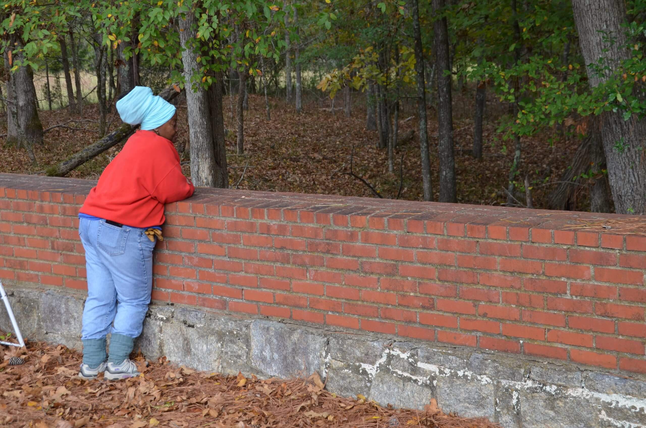 Still from Acts of Reparation. Full shot of a woman standing while leaning on a short wall in the forest. She wears a red hoodie and light blue jeans.