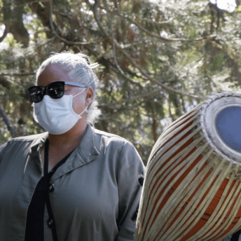 Still from Acts of Reparation. Medium shot of a woman (Selina Davidson) and a man, both of them wear sunnies and Selina wears a face mask. The man plays a big drum and has a headset microphone. It is daytime.