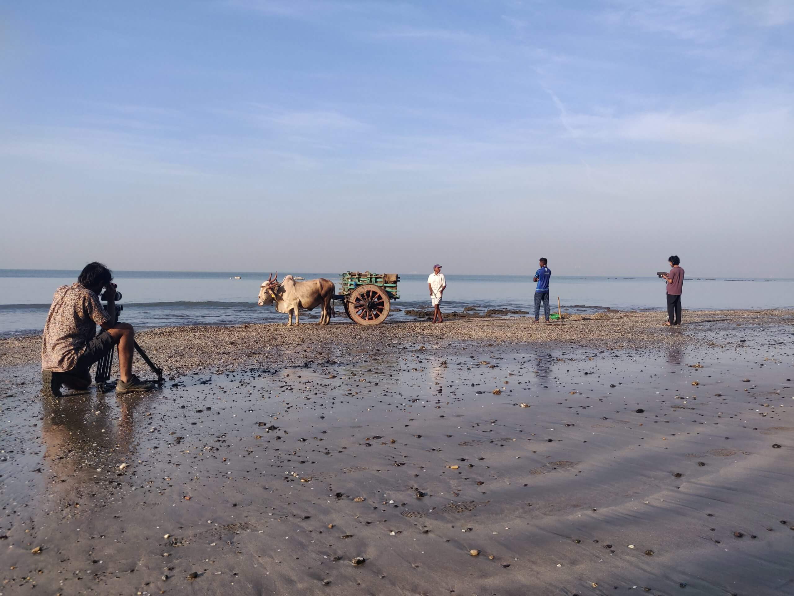 Production shot of Against the Tide. A man with a camera records other three men on the sea shore, there is a carriage with an animal pulling it and three men standing next to the carriage.