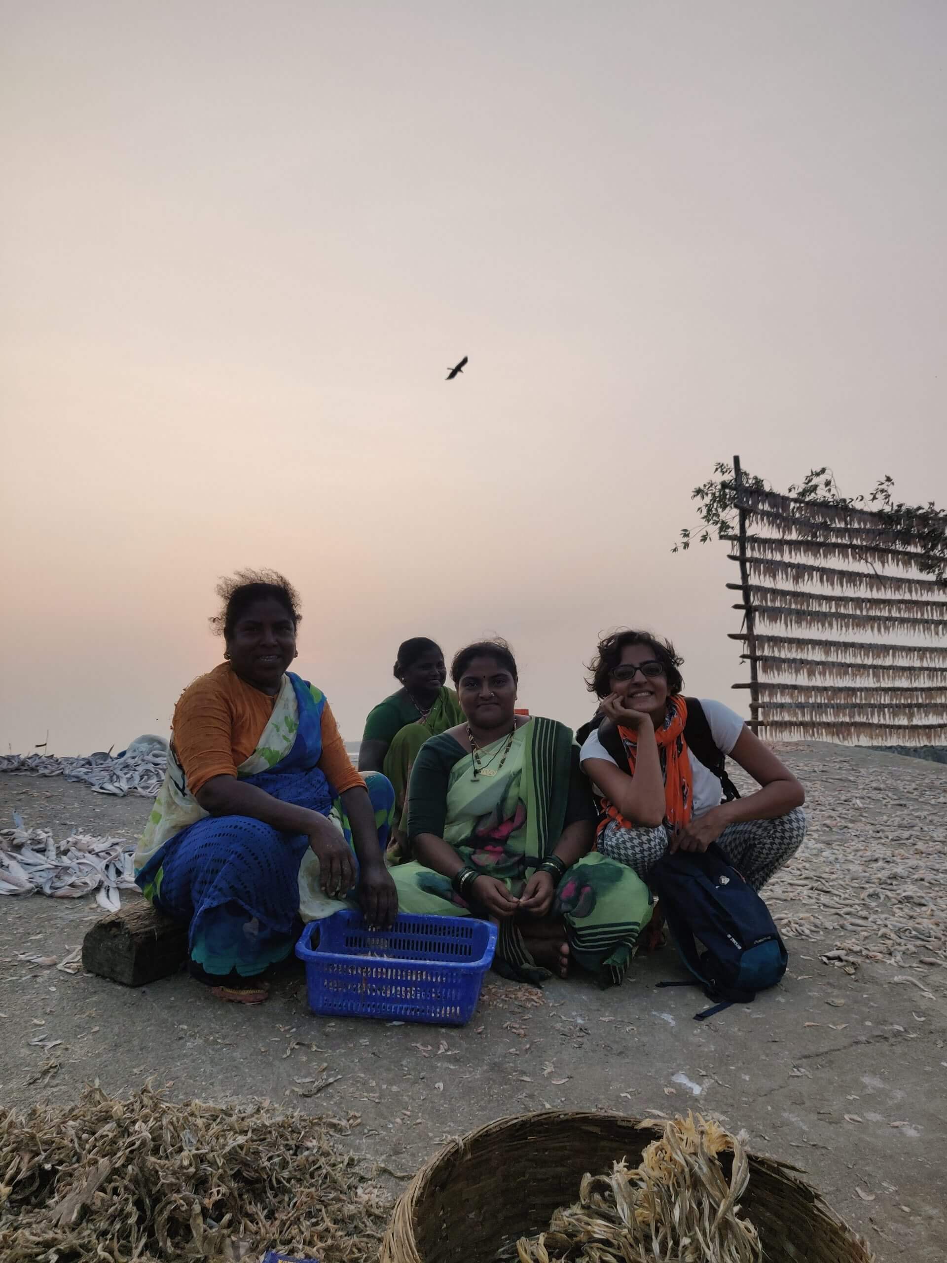 Production shot of Against the Tide. Three women sitting on the sand with baskets and with Director Sarvnik Kaur smiling.