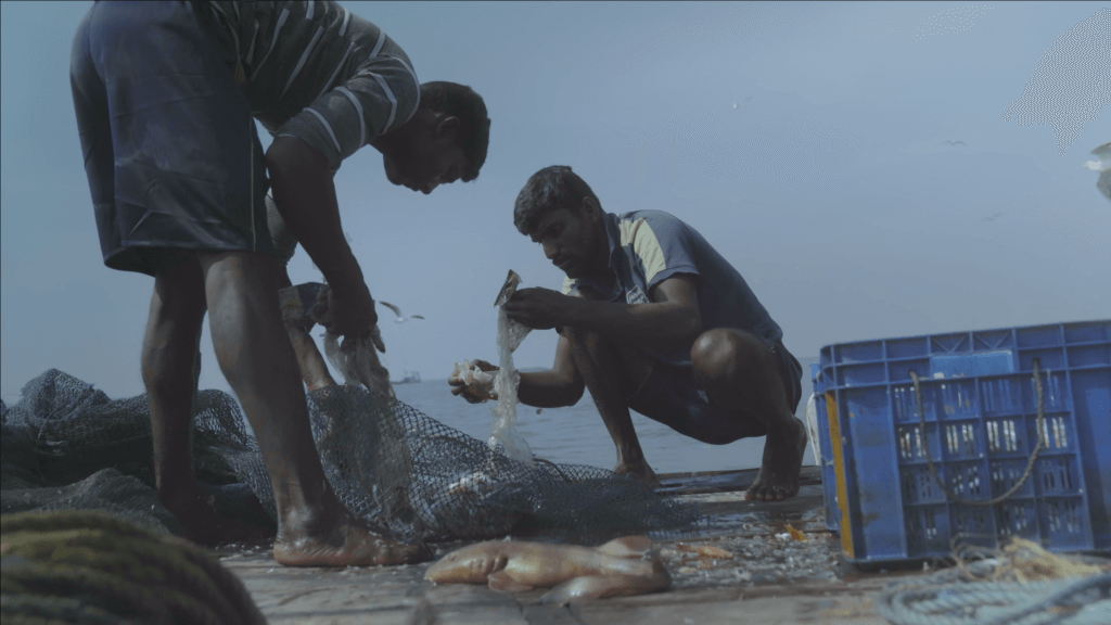 Still of Against the Tide. Two men go through the content of their fishing net, there are a fish and plastic trash.