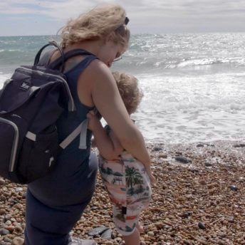 Still from Is There Anybody Out There? Ella Glednining, a blonde woman, is seen from behind, hugging her son River. They are standing on a pebbled shore with the waves in front of them.