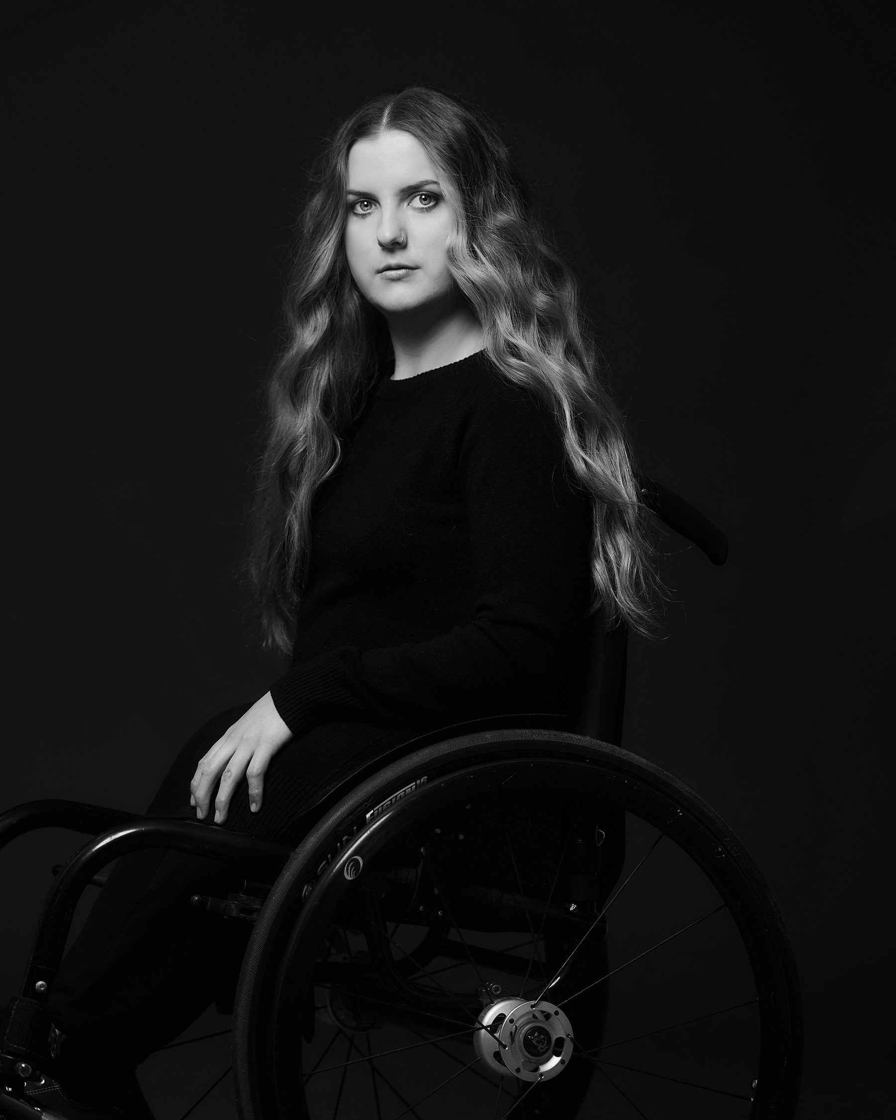 Ella Glendining looking straight ahead. She has light-colored, wavy, waist-length hair. She is wearing dark clothes and is sitting in a wheelchair. Black and white portrait.