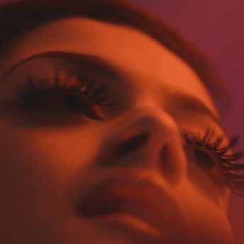 Still from Mija. An upward, close-up of a woman's face with pink/orange lighting.
