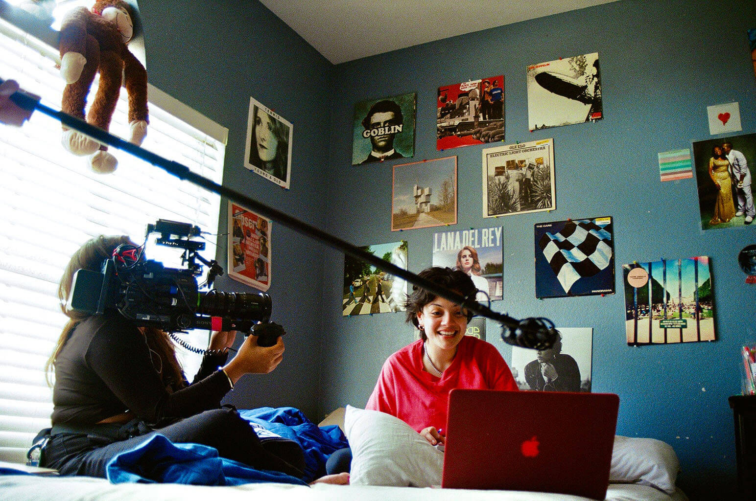 Still from Mija. A woman is sitting on her bed, smiling at her laptop. Another woman is sitting on the bed holding a camera on her shoulder, filming the other woman. There is also a boom mic in the center.