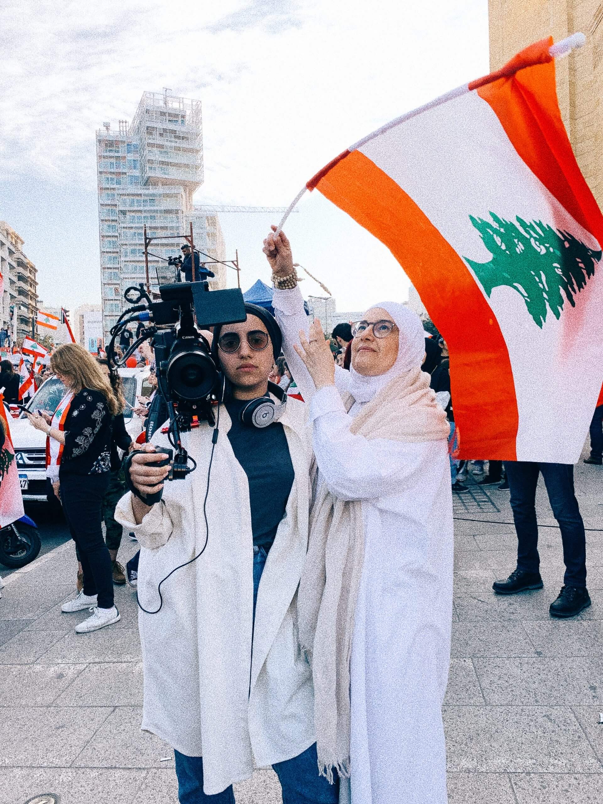 Still from Q. Filmmaker Jude Chehab is wearing sunglasses and is standing outside holding a camera over her shoulder standing next to a woman wearing glasses and a hijab, waving the flag of Lebanon, which is red and white with a green pine tree in the center.