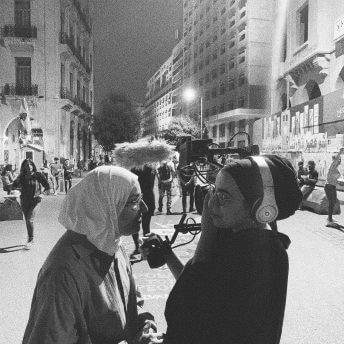 Still from Q. Two women are standing outside in a square of people. One of them is filmmaker Jude Chehab, she is wearing a headscarf and is holding a camera over her shoulder, filming the other woman wearing a hijab and glasses. Black and white processed image.