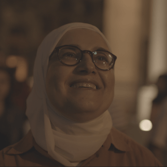 Still from Q. A woman is standing outside at night wearing glasses and a hijab, she smiles upward, away from the camera. There are other people in the background.
