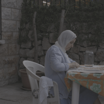 Still from Q. A woman wearing a hijab sits at a table outside. She is sorting a pile of grains into a large jar.