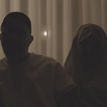 Still from Q. Two people in darkness, they are backlit. One person is wearing a hijab.
