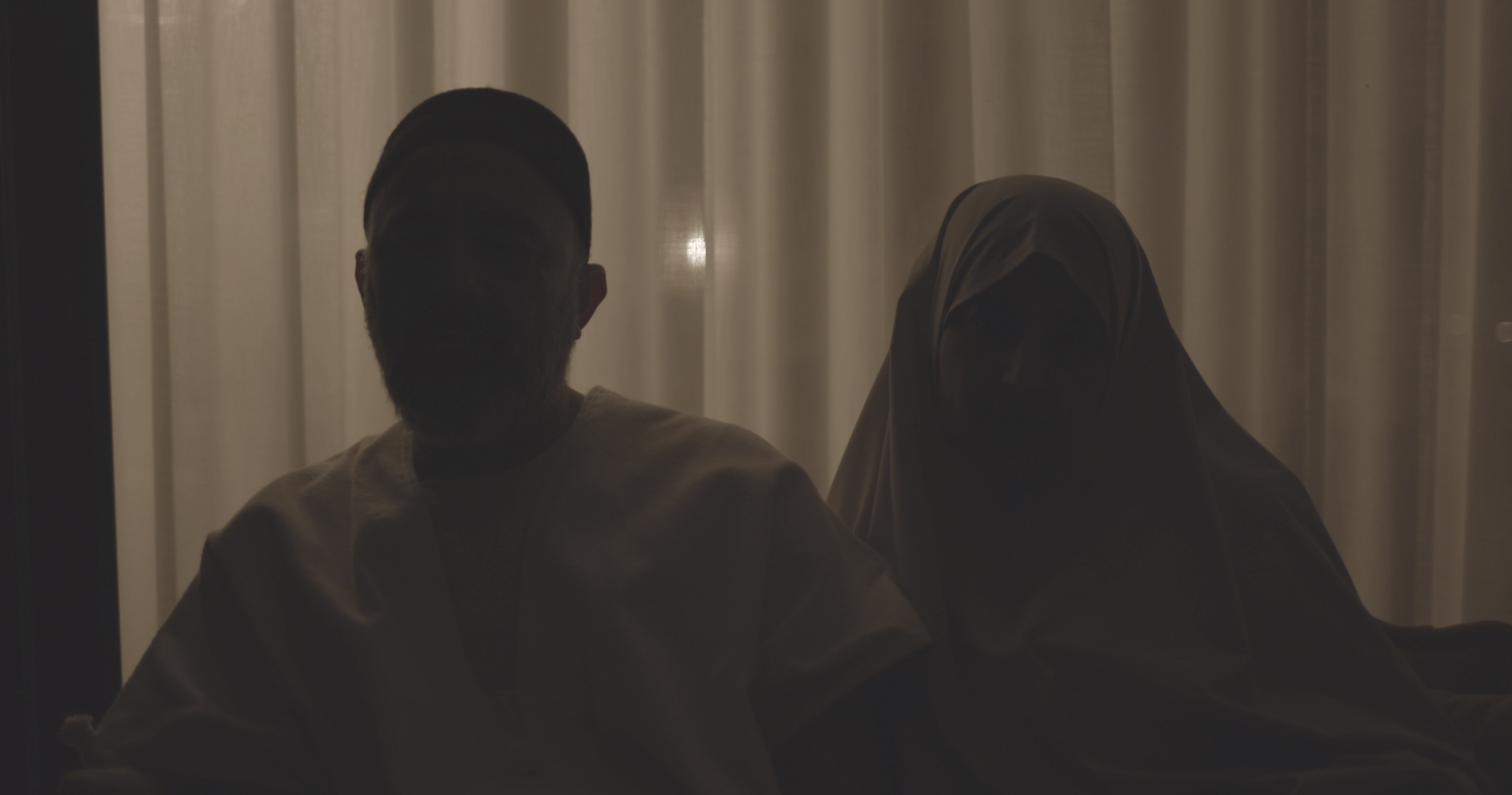 Still from Q. Two people in darkness, they are backlit. One person is wearing a hijab.