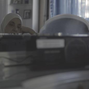 Still from Q. The reflection of a woman wearing a hijab sitting at a desk inside a bedroom with her hand on her face. The reflection of her face is blocked by a small machine sitting on the counter.